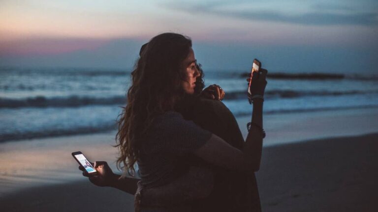 Person hugging someone by the ocean at twilight, holding two smartphones and focusing on screens, symbolizing social media’s pull
