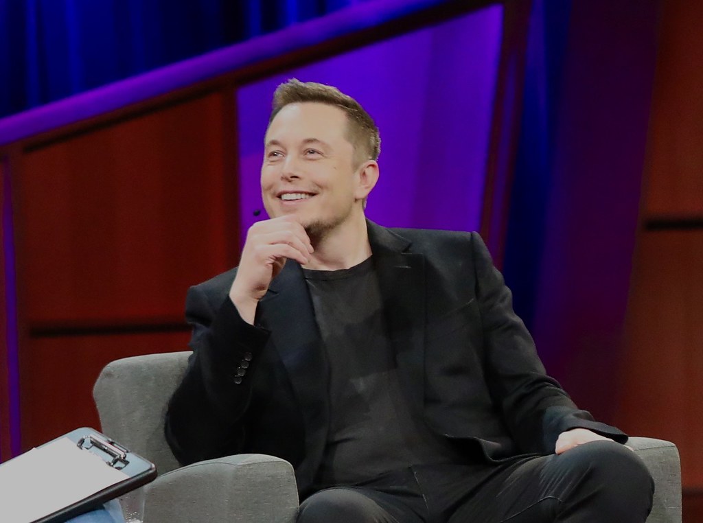 Smiling man in a black blazer sitting on a gray armchair with a purple and red stage backdrop
