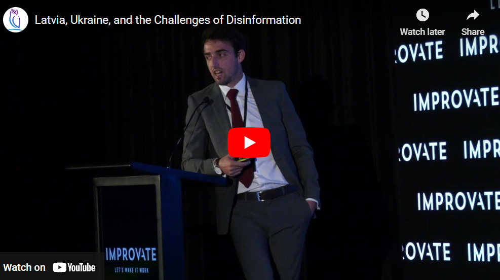 Speaker in a suit presenting at an IMPROVATE conference podium during a talk on Latvia, Ukraine, and disinformation
