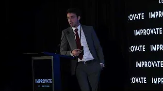 Man in suit presenting at the Improvate conference, standing beside a podium and a screen displaying the event name