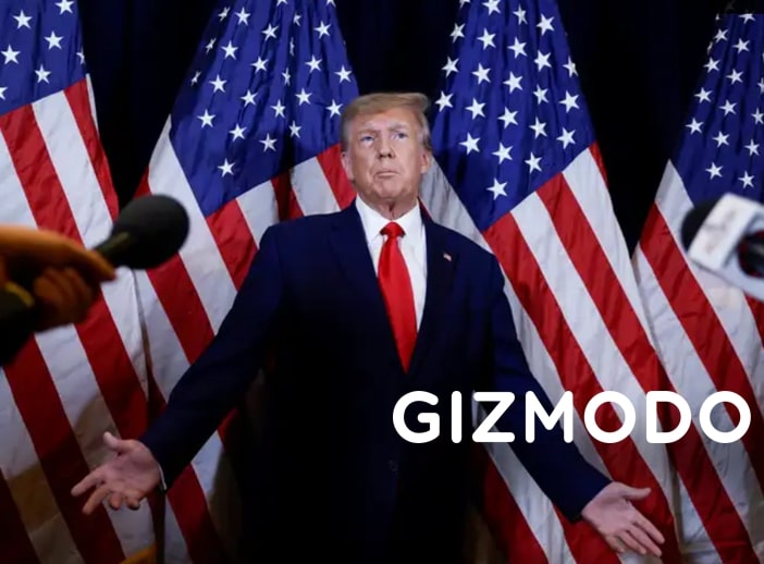 Person in dark suit and red tie stands with arms outstretched before several U.S. flags as microphones are held toward them