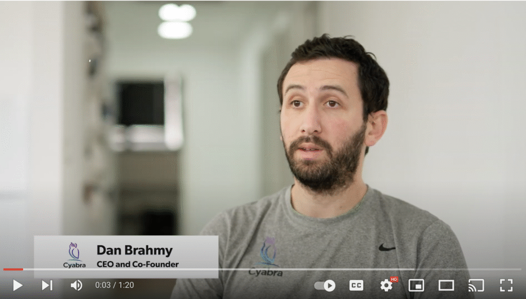 Bearded man in a gray Cyabra T-shirt speaking on camera in an office hallway, lower-third caption labeling him as CEO and co-founder