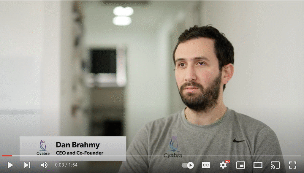 Man in gray Cyabra t-shirt during an interview, video paused at three seconds with YouTube controls visible
