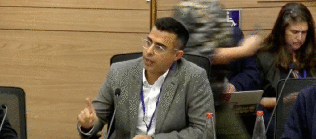 Man in glasses and grey blazer speaks into a microphone at a conference table, gesturing with one finger raised while colleagues work on laptops nearby
