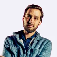 Bearded man in a blue jacket standing with arms crossed against a plain background