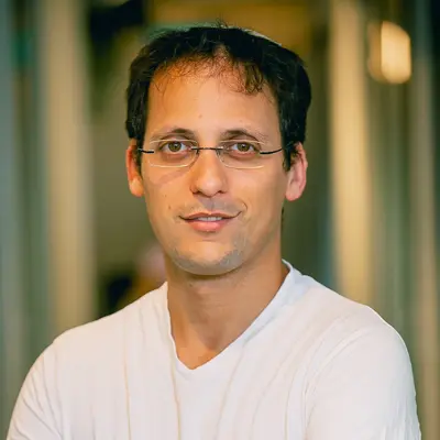 Man with short dark hair wearing glasses and a white T-shirt standing in an office hallway with blurred background