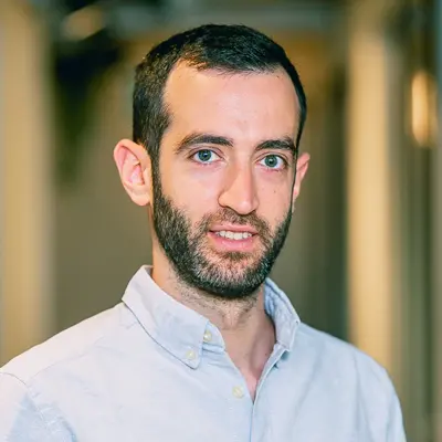 Smiling man with short dark hair and beard wearing a light blue button-down shirt in an office hallway