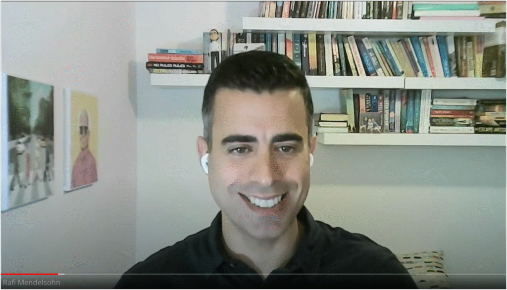 Smiling man wearing wireless earbuds during a video call with bookshelves and artwork in the background