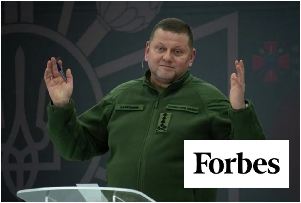 Man in green military jacket raising both hands behind a podium, Forbes logo overlay