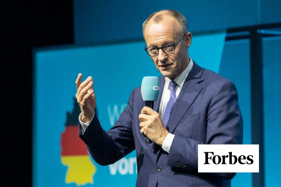Man in blue suit and glasses holding a CDU-branded microphone while speaking on stage, with a Germany silhouette graphic in the background