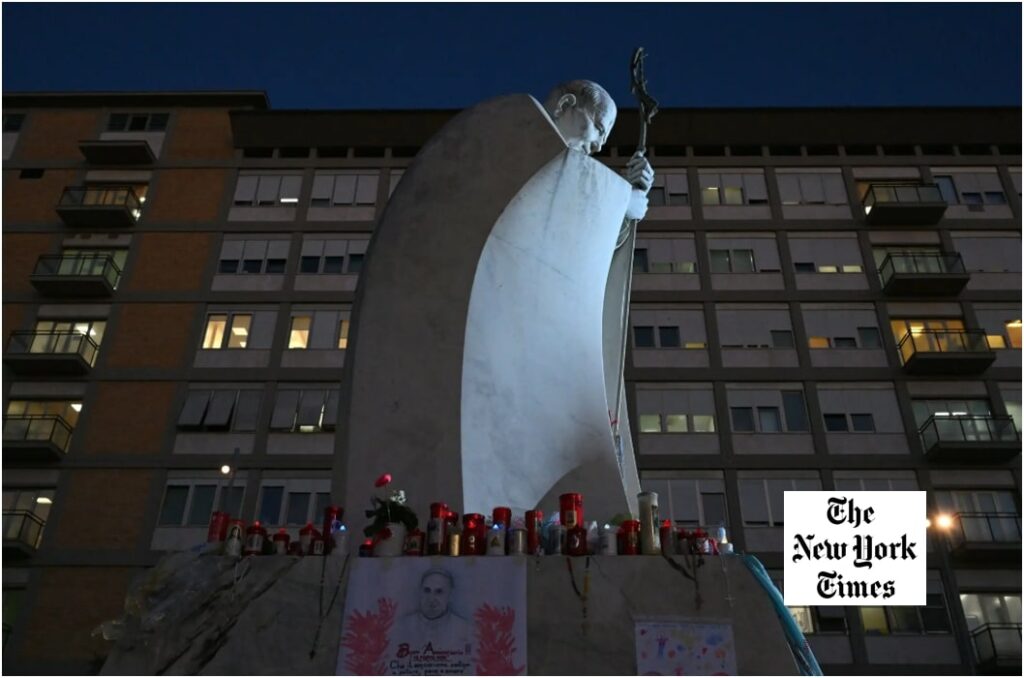 Statue of Pope John Paul II outside Gemelli Hospital in Rome at dusk, with candles, flowers and drawings placed at its base