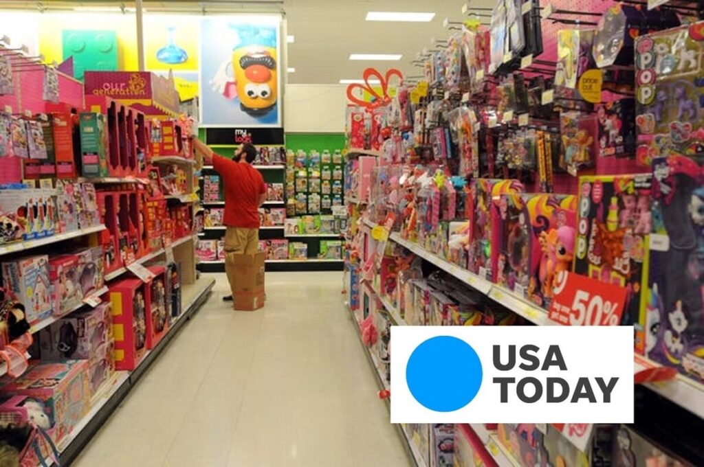 Employee restocking a brightly colored toy aisle in a big-box retail store, surrounded by shelves packed with dolls, games and 50% off sale signs