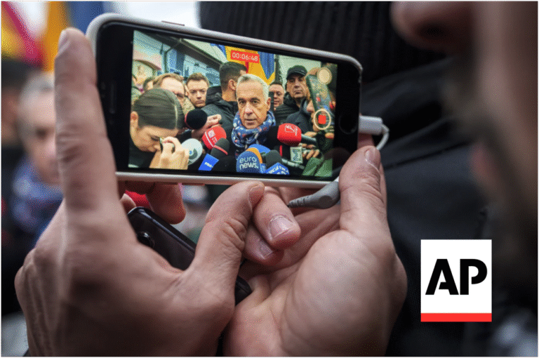 Hands holding a smartphone filming a politician surrounded by journalists and colorful microphones during a campaign media scrum
