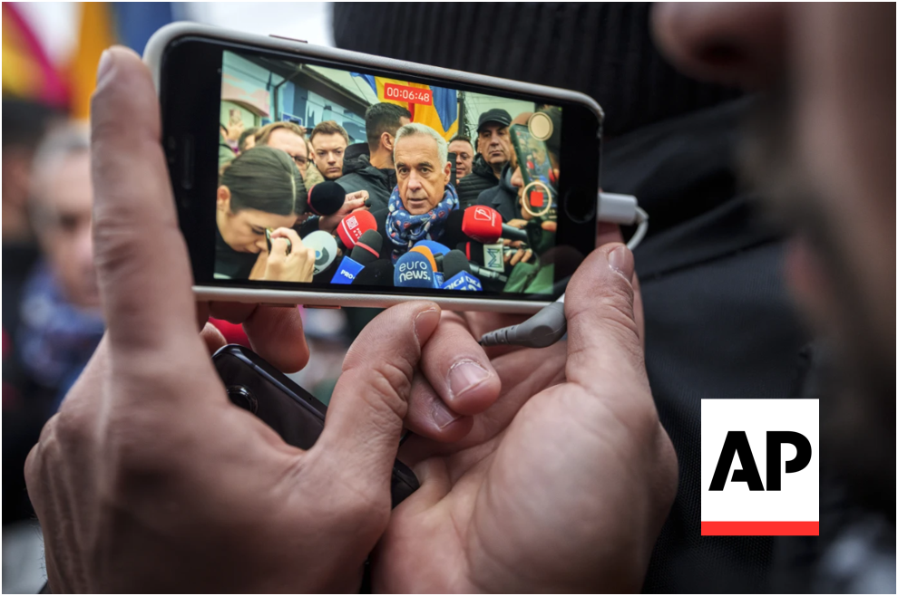 Hands holding a smartphone filming a politician surrounded by journalists and colorful microphones during a campaign media scrum