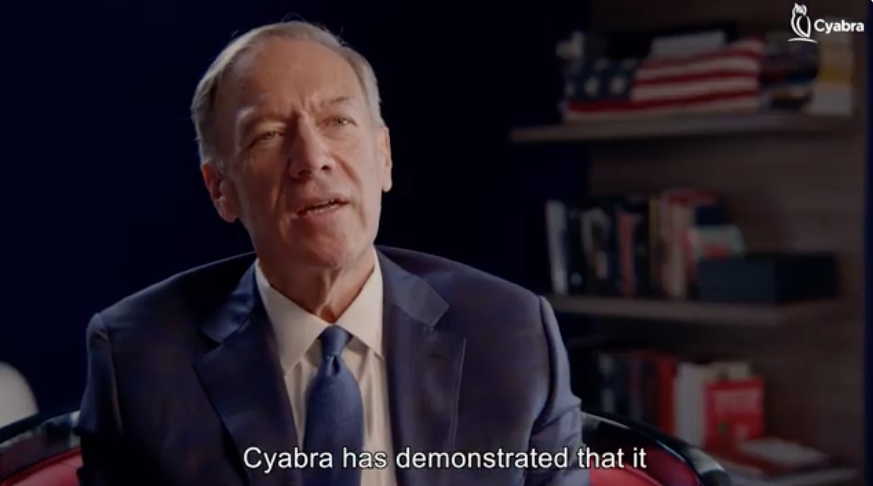 Man in a dark suit and tie speaking during an interview; bookshelf with books and a folded American flag in the background, Cyabra logo in the corner, visible subtitle reads “Cyabra has demonstrated that it”