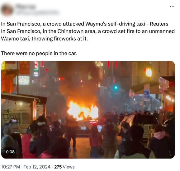 Nighttime scene in San Francisco’s Chinatown where a driverless Waymo taxi is engulfed in flames amid fireworks as a crowd gathers on the street