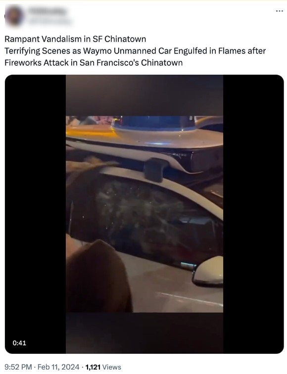 Nighttime crowd surrounds a driverless Waymo vehicle in San Francisco’s Chinatown, its windows shattered and roof-mounted sensors visible amid fire damage